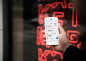 Betfair faces six-figure penalty after breaking Australian spam laws. A person holding a white smartphone displaying a messaging app filled with multiple spam messages, with a red neon sign blurred in the background.