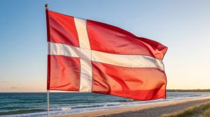 A photograph of a Danish flag waving proudly against a backdrop of a clear, sunny sky. The flag’s iconic red stripes and white Scandinavian cross ripple gently in a slight breeze, creating dynamic folds and shadows. Below the flag, a sandy beach stretches along the coastline, meeting the turquoise waters of the Baltic Sea. Warm sunlight bathes the scene, highlighting the vibrant colors of the flag and creating a cheerful atmosphere. Danish Gambling Authority updates marketing rules to protect inactive players