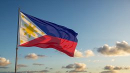 A photograph of the Philippine flag waving proudly against a clear blue sky. The flag's vibrant red and yellow colors ripple in the gentle breeze, showcasing the iconic gold sun and three yellow stars emblazoned across the blue canton. The sky is a gradient of cerulean, with scattered fluffy white clouds drifting peacefully in the distance. Sunlight bathes the scene, highlighting the flag's textures and creating a sense of national pride and serenity.