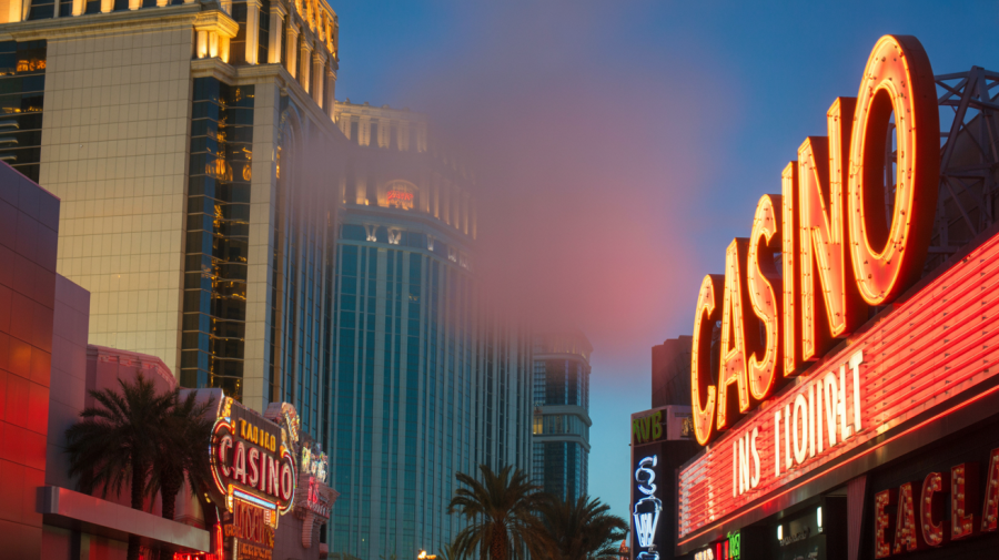 A photograph of a vibrant close-up of the Las Vegas Strip at twilight. Towering buildings rise on either side, with bright neon signs proclaiming "CASINO" in bold, stylized lettering. The buildings are a mix of gold, red, and turquoise, reflecting the dazzling city lights. A slight haze hangs in the air, softening the details and adding a touch of mystery to the iconic scene.