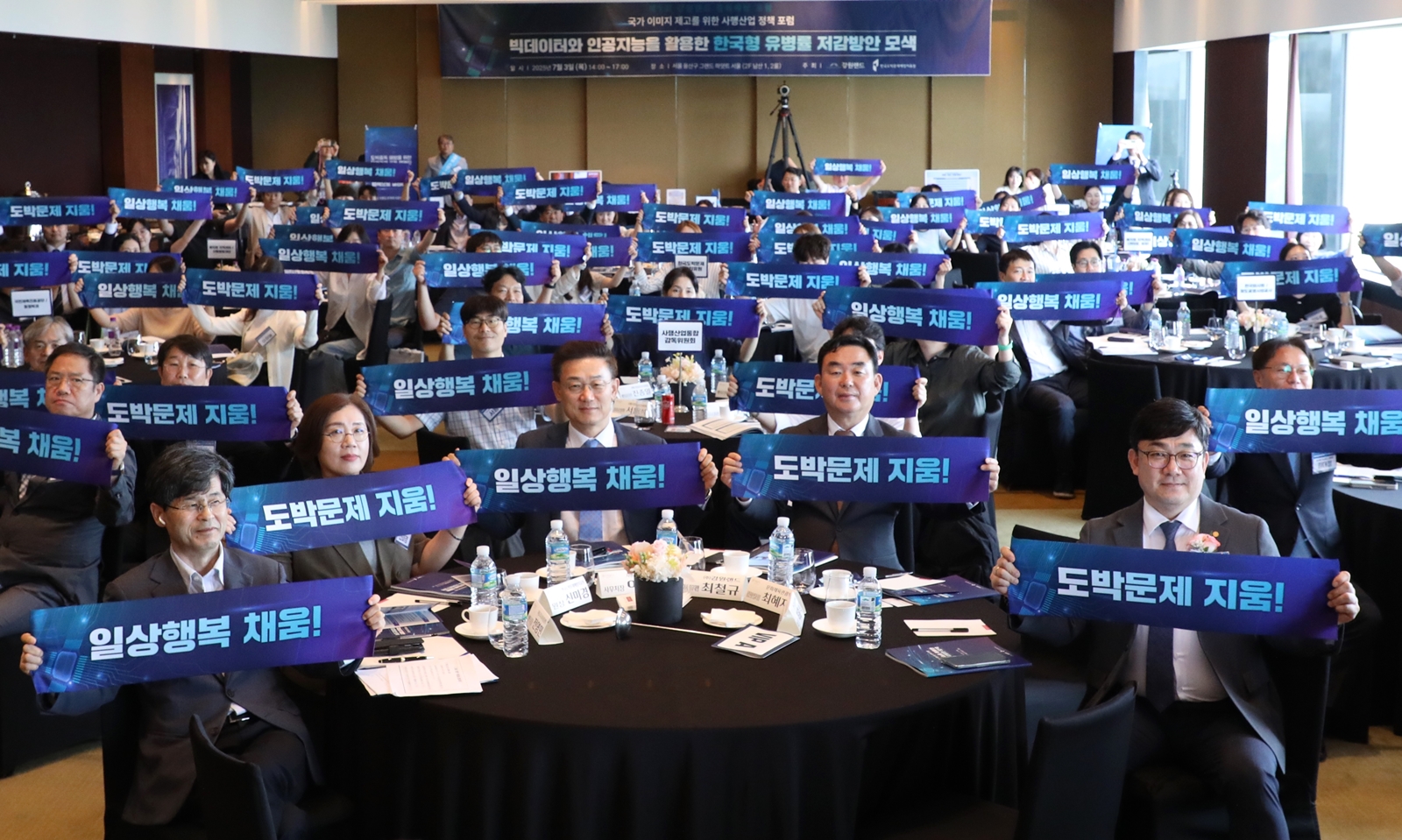 A large group of people seated at round tables in a conference room, holding up blue banners with Korean text that read “도박문제 지움!” (“Erase gambling problems!”) and “일상행복 채움!” (“Fill daily life with happiness!”). A banner above the stage mentions a forum on big data and AI in South Korea.