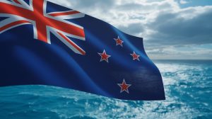 A picture that takes a close image of the New Zealand flag greatly ripples in a strong wind. The dark blue field is defined for science and four red stars sharply for a cloudy sky. Silver fern logo on science is skillfully visible. The background is a vibrant blue circumference under the spread of sunlight, which adds depth and movement to the scene.