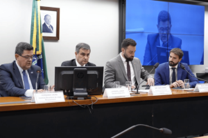 The Brazilian Congress asked to focus on the 6.4 billion dollar gambling revenue losses on the black market. A committee of four men wearing suits sitting in a long office during an official session of Congress in Brazil. Brazilian flag and a framed image hanging on the wall behind them. Paints and microphones are placed in front of each speaker, along with cups of water. A large screen in the background displays live feeding to the meeting.