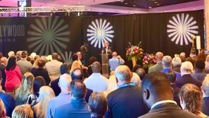 A crowd stood at the forefront of the image, in the background he could see a black curtain and a man stood on a platform in front of it. They celebrate the opening of a new casino.