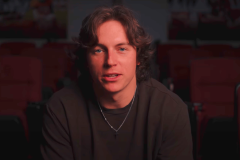 Oklahoma quarterback John Mateer has come under the spotlight after screenshots of past references to "sports gambling" surfaced on his Venmo account, but the incident has been dismissed as “inside jokes” with friends. A young man with wavy brown hair, wearing a dark sweatshirt and a silver necklace with a small cross pendant, seated indoors with red theater-style chairs in the background.