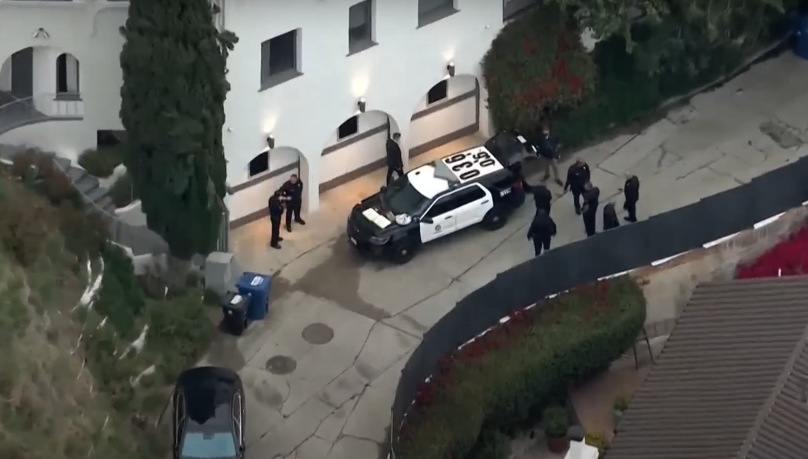 Aerial display of a multi -storey white palace with several LAPD officers and two noticeable police cars in the corridor during a clear raid or investigation. Officers are placed near the entrance to the building and along the curved corridor surrounded by green landscapes.