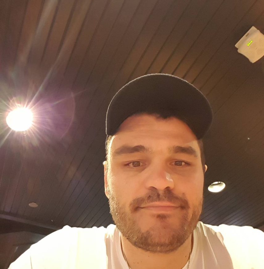 A man wears a black bispul hat and a white shirt, looking at the camera under warm internal lighting with a wooden ceiling above.