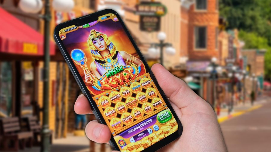 South Dakota gaming commission warns of uptick in illegal online casinos. A person holding a smartphone displaying an Egyptian-themed online slot game, with a small-town street scene blurred in the background.