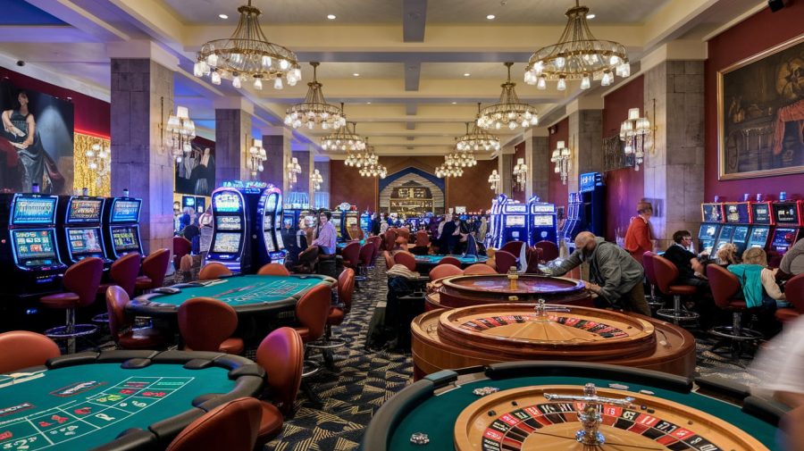 A photo of the inside of a casino with a variety of games. There are slot machines, roulette wheels, and poker tables. The walls are adorned with chandeliers and paintings. The floor is checkered. There are people playing the games. The background is dimly lit. Betting and Gaming Council strongly rejects IPPR findings on gambling tax hike