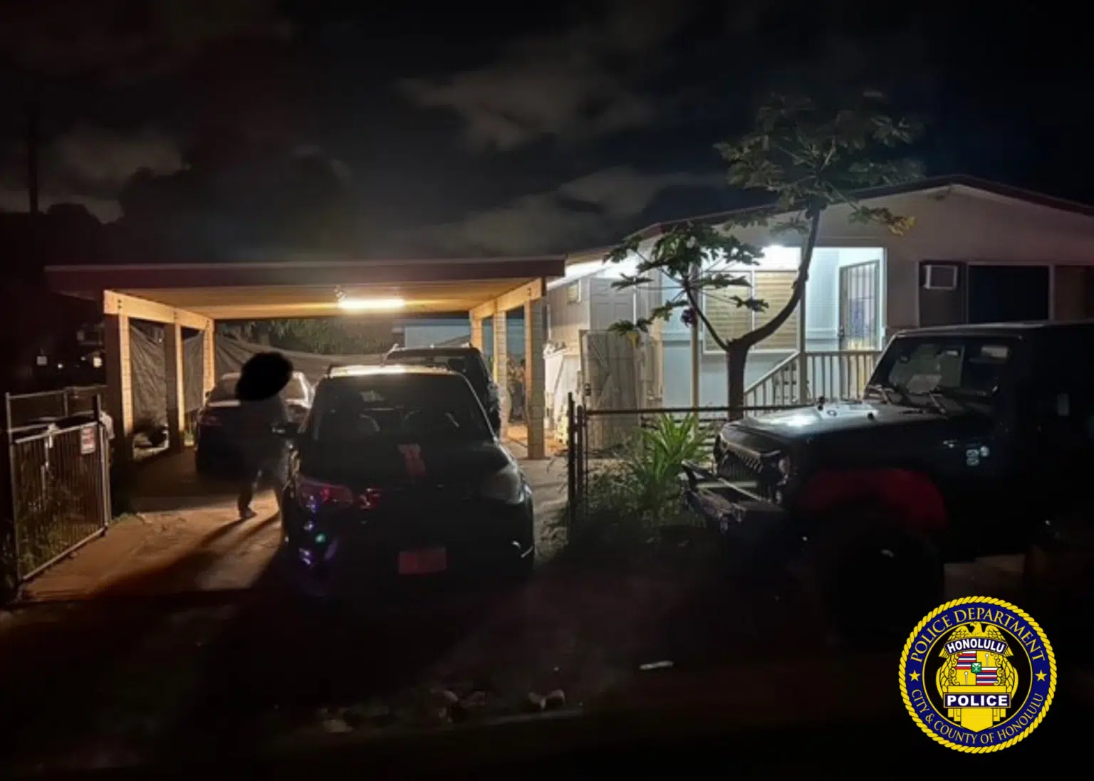 A residential house at night with cars parked in the corridor and a person walking under a garage. The house is lit from the inside, and the Honolulu Police Department logo is displayed in the bottom corner.