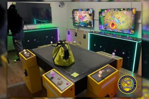 A room has many bright -style gambling machines, each with multiple controls and large screens showing games of colorful fish. A yellow bag is located on the central table, and the machines are glowed with neon lamps. Honolulu Police Department logo in the corner.