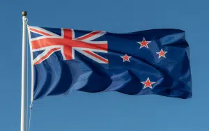 A photograph of the New Zealand flag billowing dramatically in a strong wind against a clear blue sky. The flag displays its distinctive design with the Union Jack in the upper left corner and four red stars outlined in white representing the Southern Cross constellation on a deep blue field. The fabric ripples and waves create dynamic shadows and highlights across the surface, emphasizing the flag's movement and patriotic symbolism. A tall white flagpole extends upward, with the flag attached by metal rings that catch occasional glints of sunlight as they move with the wind's rhythm.