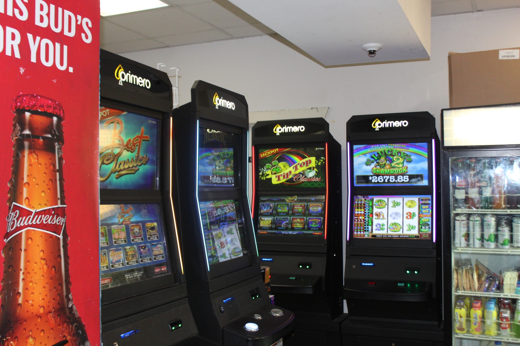 Several electronic gaming machines are lined inside a small store next to beverage coolers and Budweiser advertisement.