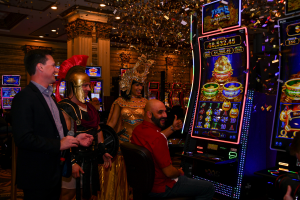 Man sat at a slot machine, in a casino, with people surrounding him. Caesars Entertainment and AGS unveil exclusive Egyptian-themed slots