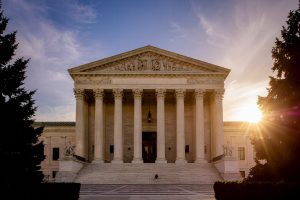 Federal Judge Sends Kalshi Vs. Massachusetts Case Back To State Court. The front facade of the U.S. Supreme Court building at sunset, showing its grand marble staircase, tall Corinthian columns, and detailed pediment sculptures. Sunlight streams in from the right side, casting a warm glow across the scene, with trees framing the image on both sides.