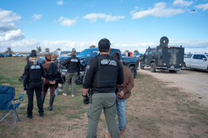 Idaho FBI raid leads to grand jury indictment of five in gambling case.Law enforcement officers wearing “State of Idaho Parole Officer” vests detain an individual during an outdoor operation. Several officers and vehicles, including an armored truck, are visible in a rural field under a partly cloudy sky.