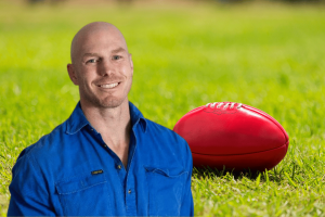 Senator David Pocock smiles, wearing a blue shirt, standing on a grass field with a red soccer ball lying on the grass behind him. Senator David Pocock has been kicked out of a sports club over a gambling sponsorship row