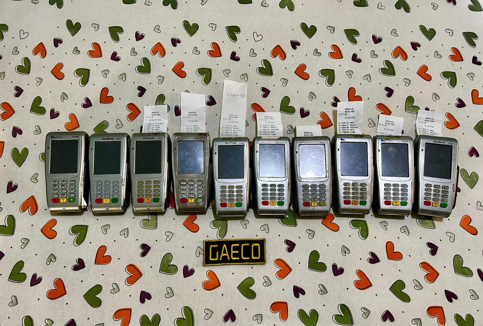 A row of seized Verifone card machines placed on a patterned tablecloth, each with receipts, with a GAECO patch displayed in front of them.