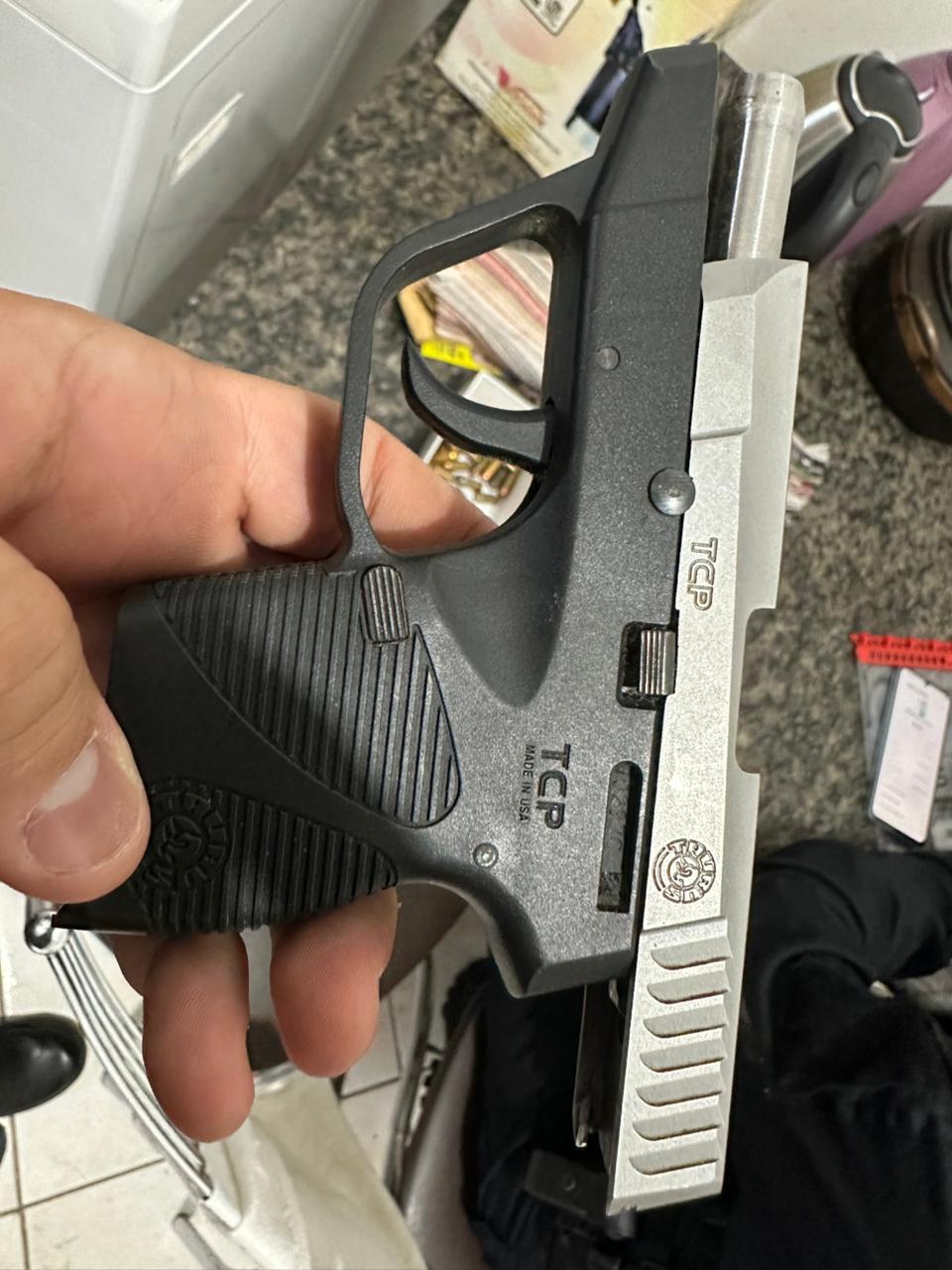 A close-up of a person holding a Taurus TCP handgun, with ammunition and other items visible in the background.