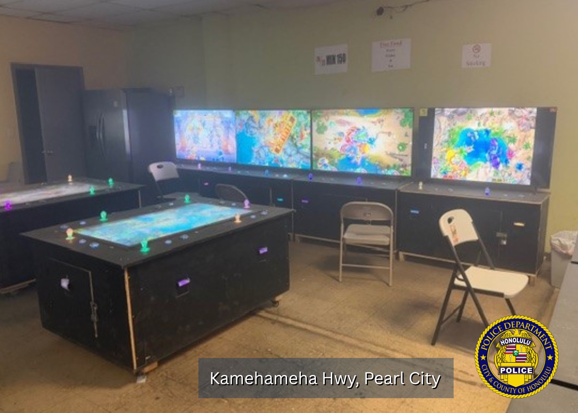 Uma sala contendo várias grandes mesas de jogos eletrônicos com telas coloridas e brilhantes e botões brilhantes, além de cadeiras dobráveis ​​espalhadas por toda parte. Vários monitores grandes montados na parede exibem gráficos de jogos animados semelhantes. Uma geladeira fica no canto de trás e cartazes simples estão pendurados na parede. Um gráfico do distintivo do Departamento de Polícia de Honolulu é sobreposto no canto inferior direito com o texto “Kamehameha Hwy, Pearl City”.