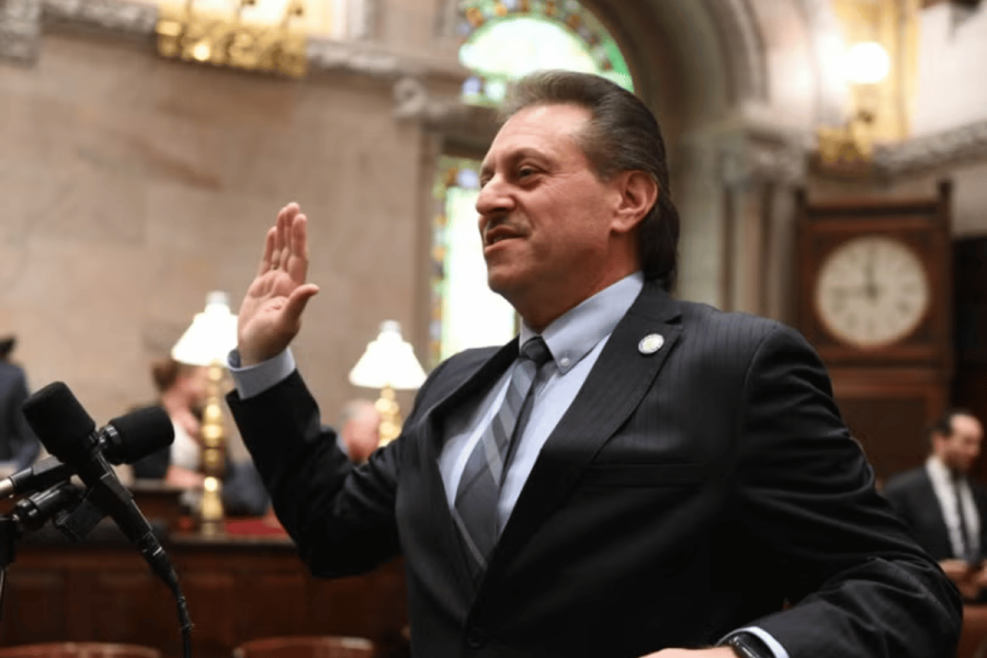 Addabbo demands swift New York regulation as prediction markets rapidly expand. Senator Addabbo in a suit raises his right hand while speaking at a microphone inside a formal government chamber with ornate décor and stained-glass windows.
