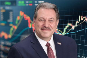 Addabbo opens New York legal review to potentially regulate prediction markets. A man in a navy suit and red patterned tie smiles in front of a background showing colorful financial charts and candlestick graphs.