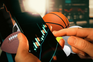 Are prediction markets gambling? Growth blurs lines between finance and betting. A person uses a smartphone displaying a candlestick trading chart while various sports balls—football, basketball, tennis ball, and golf ball—surround the device, symbolizing the overlap between financial trading and sports betting.