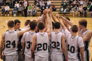 Coopersville Broncos basketball team. A high school or college basketball team huddles at center court with their hands raised together before a game. The players wear matching white jerseys with dark trim, and spectators sit in the bleachers in the background. Six student athletes found to have manipulated bets by the NCAA