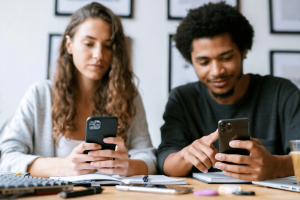 Does the betting world skew male? A woman and a man sit side by side at a desk, looking at their smartphones over open notebooks, suggesting they are comparing information or engaging in an online activity together.