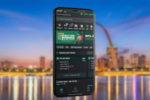 Gambling giant Caesars prepares for Missouri sports betting launch date. A smartphone displaying the Caesars Sportsbook app interface floats in front of a blurred evening cityscape. The app screen shows sports betting options, including NFL promotions, game lines, and a Thursday Night Football parlay. The background includes a riverfront skyline with bright reflections and a large arch-shaped structure.