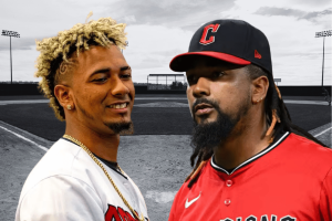 MLB players Emmanuel Clase and Luis Ortiz charged in sports betting and money laundering conspiracy. Two baseball players standing in front of a baseball field. The player on the left has short, blond curly hair and is smiling while wearing a white jersey and a gold chain. The player on the right has long dark braids, a beard, and is wearing a red baseball jersey and a black cap with a red ‘C’ on it. The background shows a grayscale baseball diamond and stadium lights.
