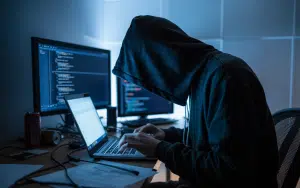 A moody photograph of a shadowy figure hunched over a glowing laptop in a dimly lit room, their face partially obscured by the blue light emanating from the screen. Connecticut warns of DraftKings scheme leading to arrests in growing fraud