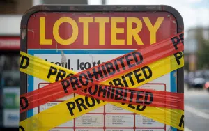 A close-up photograph of a weathered "Lottery" sign covered with strips of yellow caution tape and red "PROHIBITED" tape crisscrossing its surface. GGL warns against participating in Spanish Christmas lottery from Germany