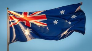 A close-up photograph of the Australian flag fluttering dramatically in a strong breeze against a clear blue sky. The bright red, white and blue fabric ripples and curves, creating dynamic shadows and highlights that emphasize the texture of the material. The constellation of the Southern Cross and the Union Jack are clearly visible as they stretch and bend with the movement of the wind, while the edges of the flag flutter abruptly in the daylight. Golden sunlight illuminates the flag from one side, creating a patriotic and dynamic composition that captures the energy of the wind.