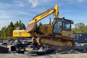Florida Gaming Control Commission destroys 147 illegal slot machines. A yellow CAT bulldozer drives over and crushes a large pile of broken electronics and metal debris on a concrete lot. Behind it, a yellow Komatsu excavator is parked near a wooded area under a blue sky.