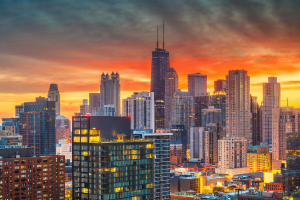 Sports Betting Alliance sues Chicago over new tax and licensing plan. Chicago skyline at sunset with illuminated skyscrapers, including the John Hancock Center, glowing against an orange and blue sky.