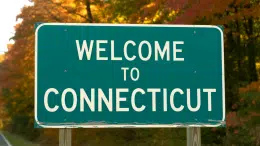 A close-up photograph of a roadside welcome sign featuring bold white lettering that reads "Welcome to Connecticut" against a deep forest green background. The sign is mounted on sturdy wooden posts and shows subtle weathering from the elements, with small chips in the paint and a thin layer of road dust along the bottom edge. Behind the sign, blurred autumn foliage in shades of amber and crimson creates a warm, inviting backdrop typical of New England's fall season. Soft afternoon sunlight filters through the trees, casting gentle shadows across the sign's surface and highlighting the crisp, official state typography.
