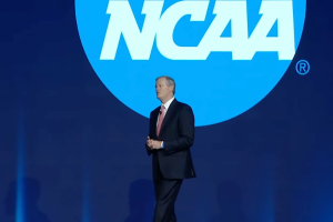NCAA logo in the background, with the NCAA President Charlie Baker stood in front of it. He's wearing a suit and giving a speech on stage to an audience. NCAA urges gambling commissions to eliminate prop bets