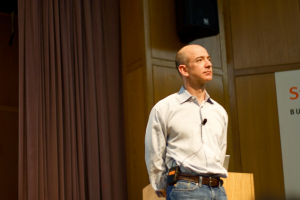 Jeff Bezos, founder of Amazon, stood on a stage looking out to the audience. Close-up of him, he's weating a light colored shirt, belt, and jeans. His hands are behind his back. Amazon founder Jeff Bezos denies Polymarket post made about his alleged advice