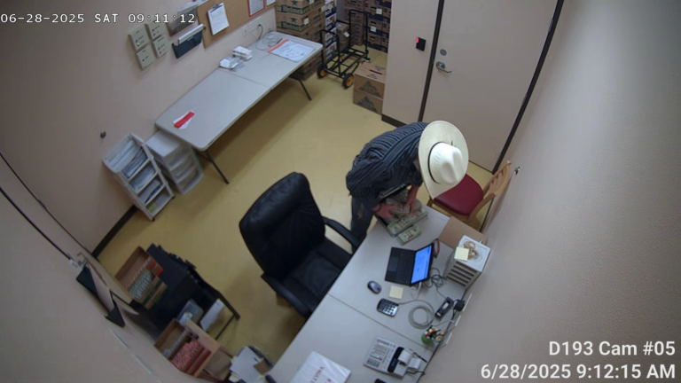 Visão de vigilância aérea de um homem de chapéu branco contando dinheiro em uma mesa dentro de um escritório com caixas e papelada.