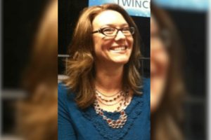 The treasurer of Minnesota youth softball is accused of stealing $110,000 for casino games. Smiling woman with glasses wearing a blue top and layered necklace, photographed indoors against a blurred background.