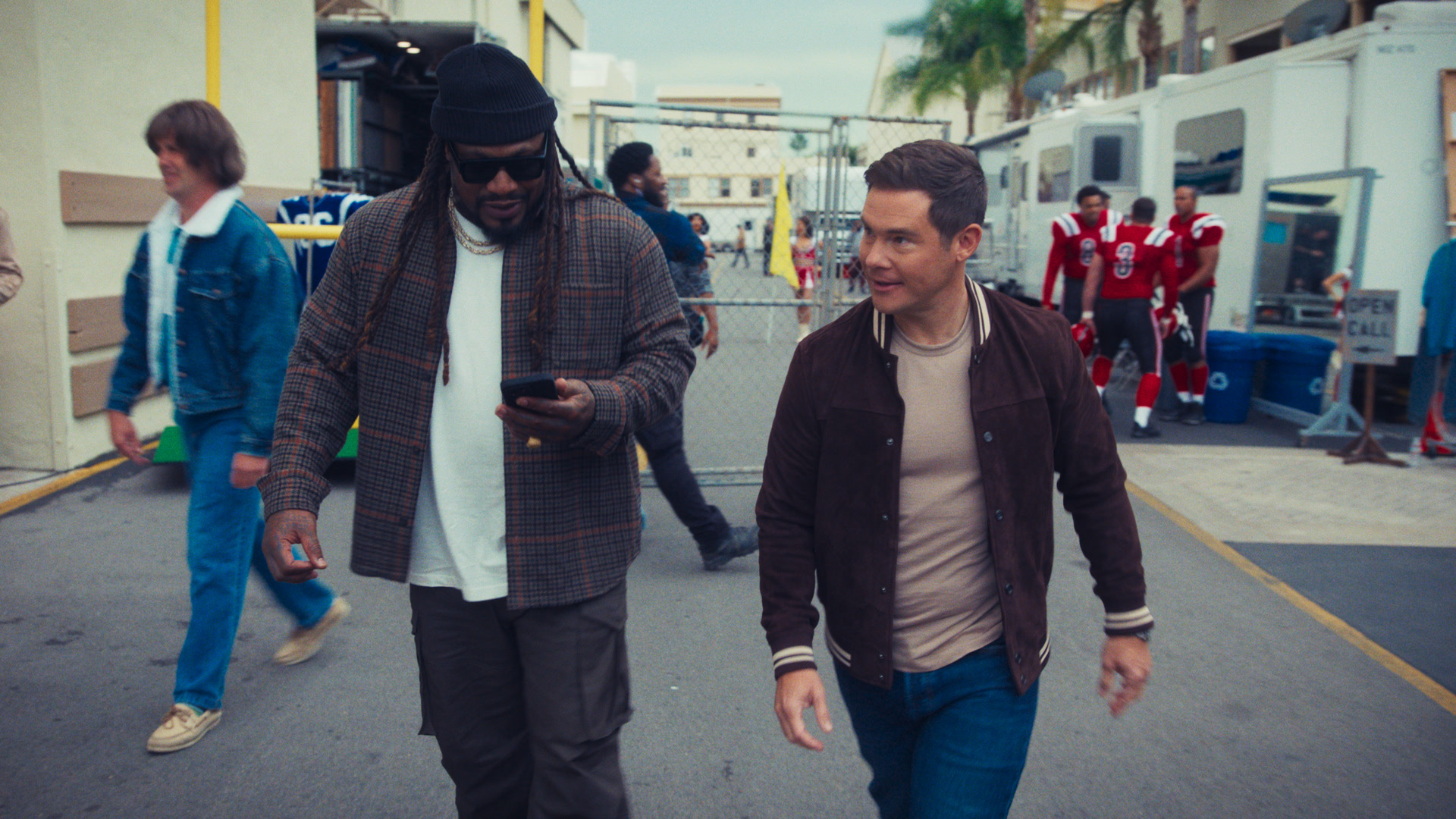 Marshawn Lynch e Adam Devine caminham lado a lado em um set de filmagem que lembra o saguão de um estádio de futebol, com jogadores em uniformes vermelhos e trailers da equipe visíveis atrás deles.