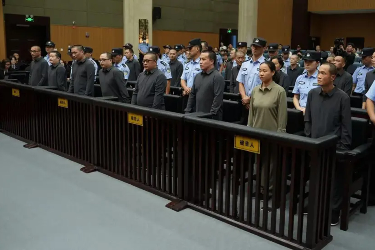 A courtroom scene showing a large group of defendants standing behind dark wooden bars marked with