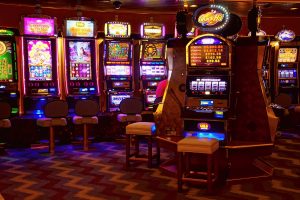 Over $3M allegedly gained in fraudulent gambling grant funds. Row of brightly lit casino slot machines inside a gambling venue, with stools positioned in front of each machine and colorful digital displays glowing in a dimly lit room.