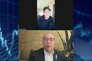 Congress knows prediction markets violate rules but won’t act without cover, says IGA chair. Screenshot of a split-screen video webcast featuring Victor Rocha on the top and Chairman David Z. Bean on the bottom. Rocha appears wearing a dark baseball cap, glasses, and a dark jacket, speaking from a home office setting. Bean appears in a suit jacket and white shirt with a lapel pin, seated in front of a textured wall with plants behind him. A blue financial chart graphic borders the sides of the image.