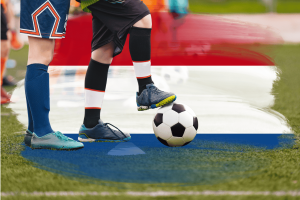 The Dutch regulator has flagged One Casino and bet365 for illegal soccer betting. The lower legs and feet of two young soccer players on a grass field, one foot resting on a black-and-white ball, with a blurred red, white, and blue background suggesting the Netherlands.