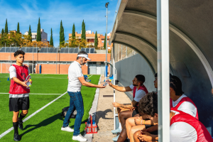 French gambling regulator ANJ warns on World Cup betting ads surge concerns. A soccer coach hands out water bottles to players wearing red training bibs as they sit on the bench during a break on a sunny outdoor field, with one player standing nearby holding additional bottles.