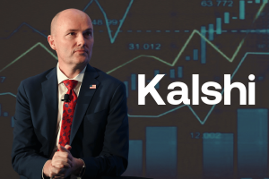 A bald man in a navy suit and red patterned tie sits with his hands clasped, wearing an American flag lapel pin and a microphone, in front of a large screen displaying financial charts and the word “Kalshi.” Kalshi files lawsuit against Utah officials over threat to prediction markets