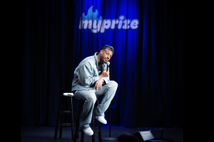 Marlon Wayans in denim jacket and backward cap sits on a stool holding a microphone on a blue-lit stage with “myprize” projected behind him.
