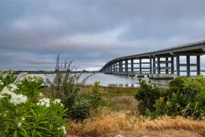 Land with a bridge-like structure on the right side and water flowing underneath. Shrub greenery is in the foreground of the image, with the sky dark blue and cloudy.